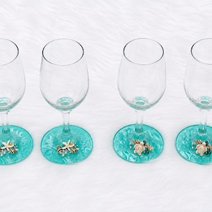 May include: Four clear wine glasses with turquoise bases. Each base is decorated with seashells and beads. The glasses are arranged on a white, textured surface.