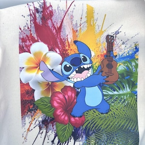 May include: Cream tote bag with a blue cartoon character holding a ukulele. The design includes vibrant paint splashes, tropical flowers, and palm leaves. The character has large eyes and a wide grin.