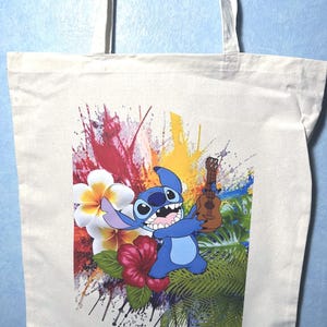 May include: A cream tote bag with a colourful graphic of the character Stitch from Lilo & Stitch. Stitch is blue and holding a ukulele. The background includes tropical flowers and paint splatters.