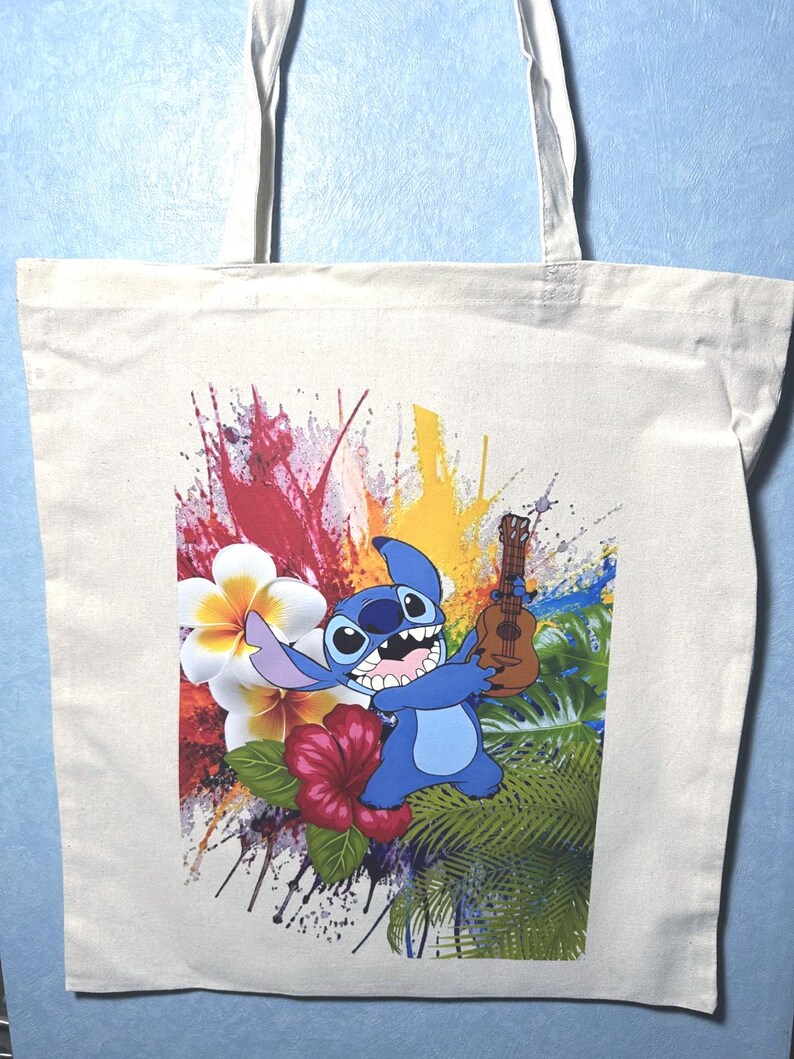 May include: A cream tote bag with a colourful graphic of the character Stitch from Lilo & Stitch. Stitch is blue and holding a ukulele. The background includes tropical flowers and paint splatters.