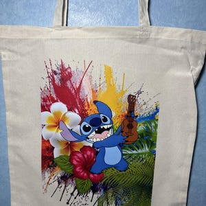 May include: A beige canvas tote bag with a vibrant print of a blue cartoon character playing a ukulele. The design incorporates tropical flowers and colourful paint splashes in red, yellow, and blue.