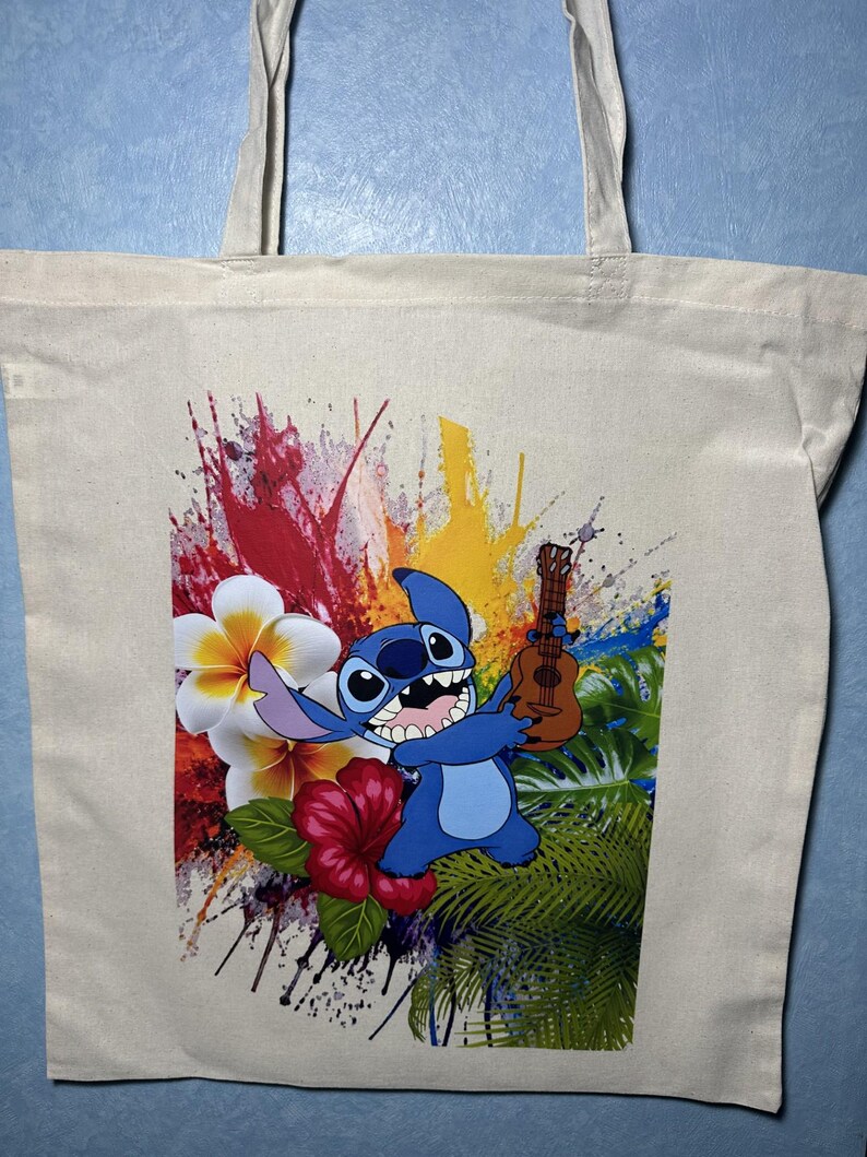 May include: A beige canvas tote bag with a vibrant print of a blue cartoon character playing a ukulele. The design incorporates tropical flowers and colourful paint splashes in red, yellow, and blue.