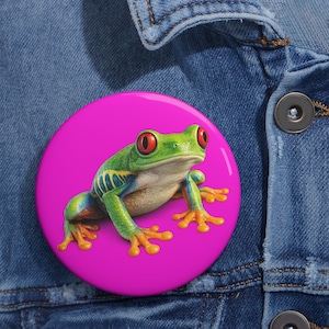 Op de afbeelding: Een ronde pin-back button met een levendige roodoogmakikikker met groene huid, blauwe en gele zijmarkeringen en oranje voeten, geplaatst tegen een effen felroze achtergrond, bevestigd aan een kledingstuk van blauwe denimstof.