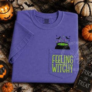 May include: A purple short-sleeve t-shirt with a chest pocket. The pocket features an embroidered black cauldron with green liquid, purple bubbles, and black bats. The text "FEELING WITCHY" is embroidered in lime green on the pocket front.
