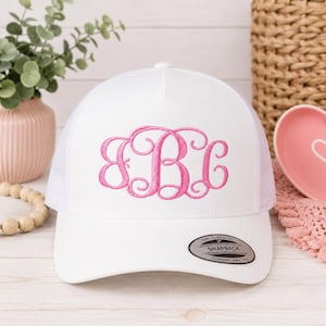 May include: White trucker hat with a pink embroidered monogram. The hat features a white front panel with a pink script monogram and a white mesh back. The hat has a curved brim and a snapback closure. The hat is a classic accessory.