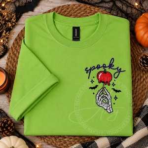 May include: A lime green short-sleeve t-shirt featuring an embroidered design of a skeleton hand holding a red candy apple. The word spooky is embroidered in purple cursive script above the apple, accompanied by small black bat and star icons.