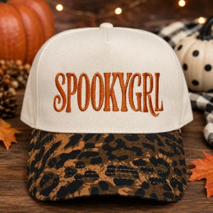 May include: A cream-colored baseball cap featuring the text SPOOKYGRL embroidered in orange thread across the front panel. The cap has a curved brim with a brown and black leopard print pattern and a button on the top center of the crown.