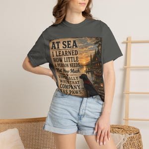 May include: A charcoal gray t-shirt featuring a graphic print of a sunset lake scene with a cabin and a black dog wearing a red bandana. The text reads: AT SEA I LEARNED HOW LITTLE A PERSON NEEDS, Not How Much. ESPECIALLY WHEN THAT COMPANY HAS PAWS.