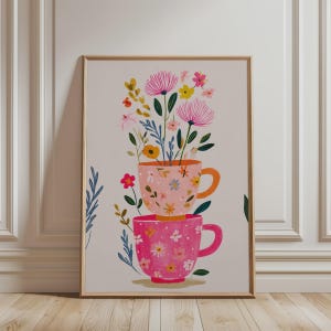 Op de afbeelding: Een ingelijste kunstdruk met een verticale stapel van twee theekopjes met bloemenpatroon in lichtroze en donkerroze. Een kleurrijk boeket van diverse bloemen en groene bladeren komt uit de bovenste kop, met extra gebladerte rondom de kopjes.