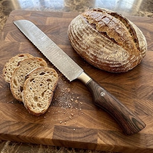 May include: A serrated bread knife with a metallic blade and a dark brown wood grain handle, positioned next to a whole loaf of sourdough bread and three sliced pieces on a wooden cutting board with a visible grid pattern of wood blocks.