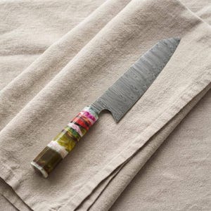 May include: A kitchen knife featuring a silver-toned blade with a visible wavy Damascus steel pattern and a cylindrical handle crafted from multi-colored resin in shades of green, pink, red, and white with a dark brown base section.