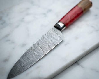 May include: A kitchen knife featuring a sharp, pointed blade with a distinct wavy Damascus steel pattern. The handle is constructed from a combination of polished wood at the end and a vibrant, translucent red resin section near the silver bolster.