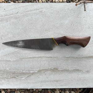 Puede incluir: Un cuchillo de chef de acero de Damasco con mango de madera oscura y refuerzo de latón. La hoja tiene un patrón distintivo y el mango tiene forma ergonómica. El cuchillo está diseñado para uso en la cocina.