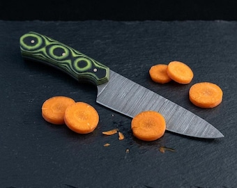 May include: A fixed-blade knife with a textured, layered green and black handle and a metallic blade featuring a fine, linear pattern. The knife is positioned alongside several round, orange carrot slices on a dark, textured surface.