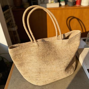 Può includere: Una borsa tote beige intrecciata con due manici arrotondati. La borsa ha un'ampia apertura e una superficie strutturata. La borsa è realizzata in un materiale di fibra naturale e si trova su una superficie grigia.