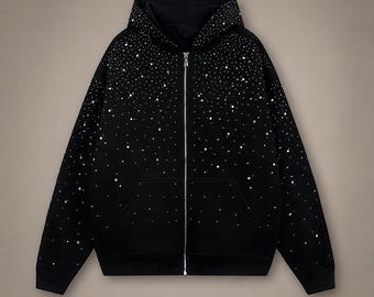 May include: A black zip-up hoodie featuring a silver metallic zipper and a dense pattern of scattered white rhinestones that are concentrated heavily around the hood and shoulders, gradually thinning out toward the bottom hem and sleeves.