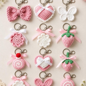 Op de afbeelding: Een collectie van twaalf gehaakte sleutelhangers met diverse vormen, waaronder roze strikken, wit-roze harten, witte vlinders, roze-witte bloemen, roze aardbeien met groene topjes en roze-witte cirkelvormige patronen met een spiraal en lintjes.