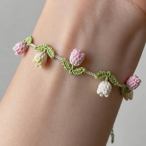 May include: A delicate crochet bracelet featuring a series of small, three-dimensional tulip flowers in soft shades of pastel pink and cream, interspersed with intricate green crocheted leaves on a thin, white thread base.