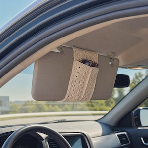 May include: A beige crochet car visor organizer sleeve featuring a textured star-like stitch pattern. The sleeve is wrapped around a gray car sun visor and contains a pair of dark-framed sunglasses tucked into the open pocket compartment.