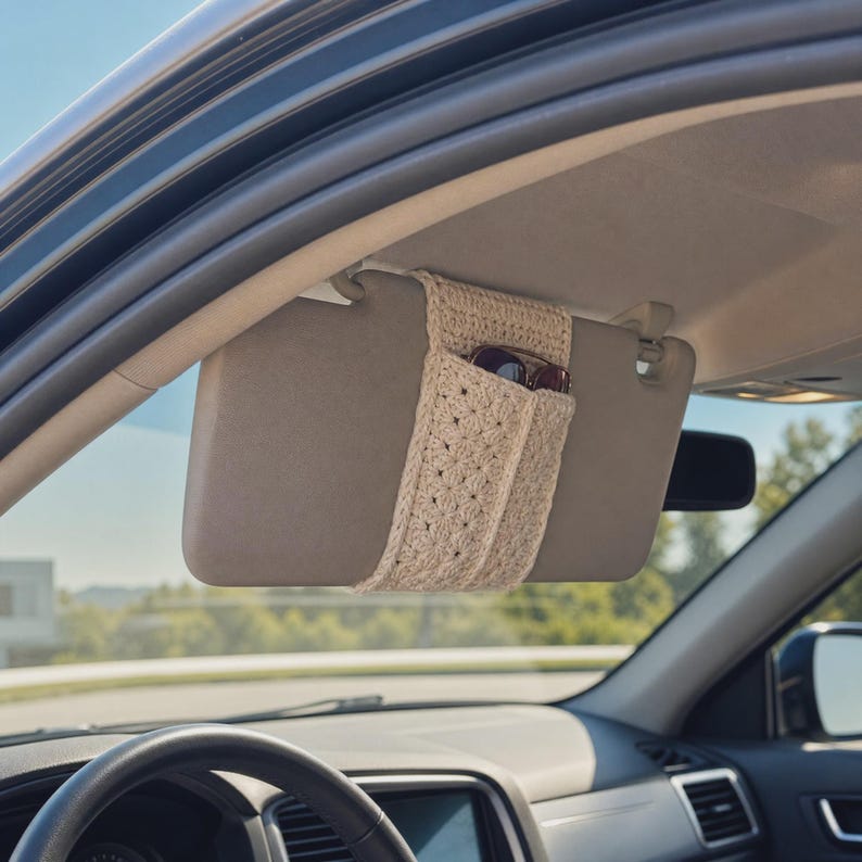 May include: A beige crochet car visor organizer sleeve featuring a textured star-like stitch pattern. The sleeve is wrapped around a gray car sun visor and contains a pair of dark-framed sunglasses tucked into the open pocket compartment.