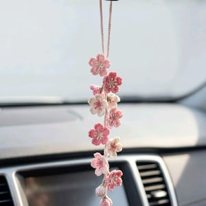 May include: A hanging car charm featuring a vertical arrangement of crocheted cherry blossom flowers in various shades of pink and white. The floral strand is suspended by a thin, light pink cord and includes several open blossoms and small buds.