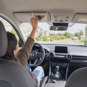 May include: A cream-colored, rectangular crochet visor organizer attached to the driver-side sun visor of a car. The organizer features a textured, open-weave pattern and is designed to hold small items like sunglasses securely against the visor.