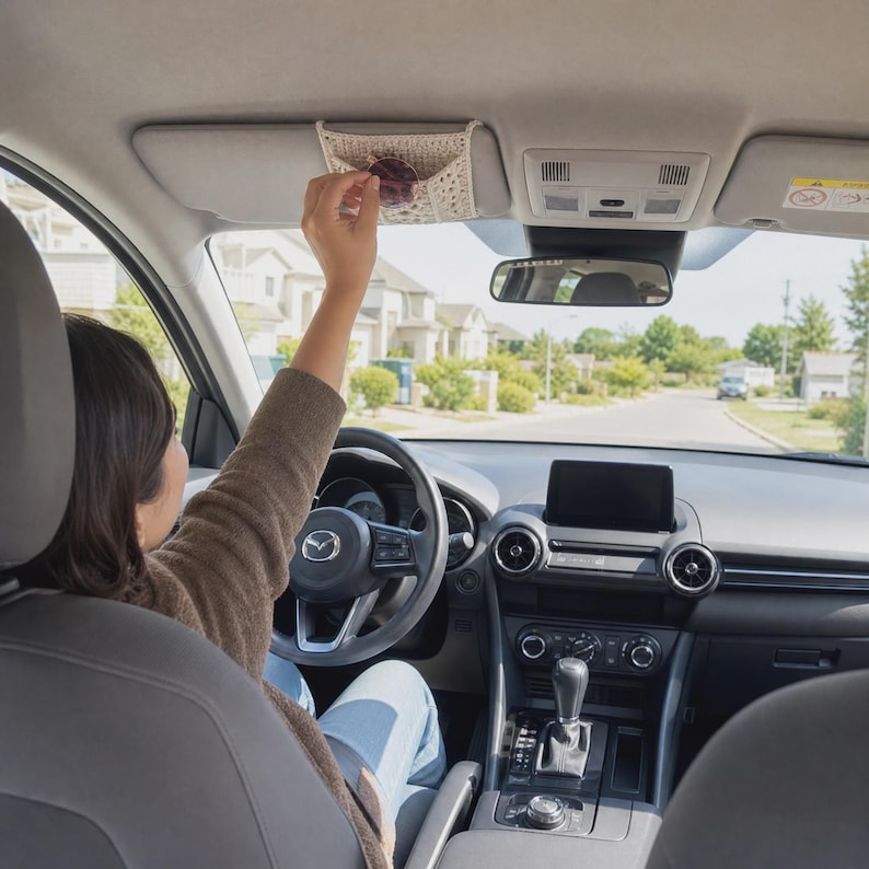 May include: A cream-colored, rectangular crochet visor organizer attached to the driver-side sun visor of a car. The organizer features a textured, open-weave pattern and is designed to hold small items like sunglasses securely against the visor.