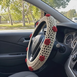 May include: A beige crochet steering wheel cover featuring a lattice pattern and small, evenly spaced red crocheted flower appliqués attached to the outer edge. The cover is fitted onto a black steering wheel inside a vehicle interior.