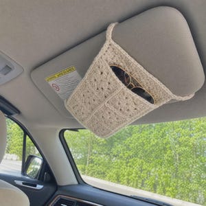 May include: A beige crochet car visor organizer featuring a textured square pattern. The accessory is attached to a vehicle sun visor and contains a pair of dark-rimmed sunglasses tucked into the open pocket, providing storage for small items.