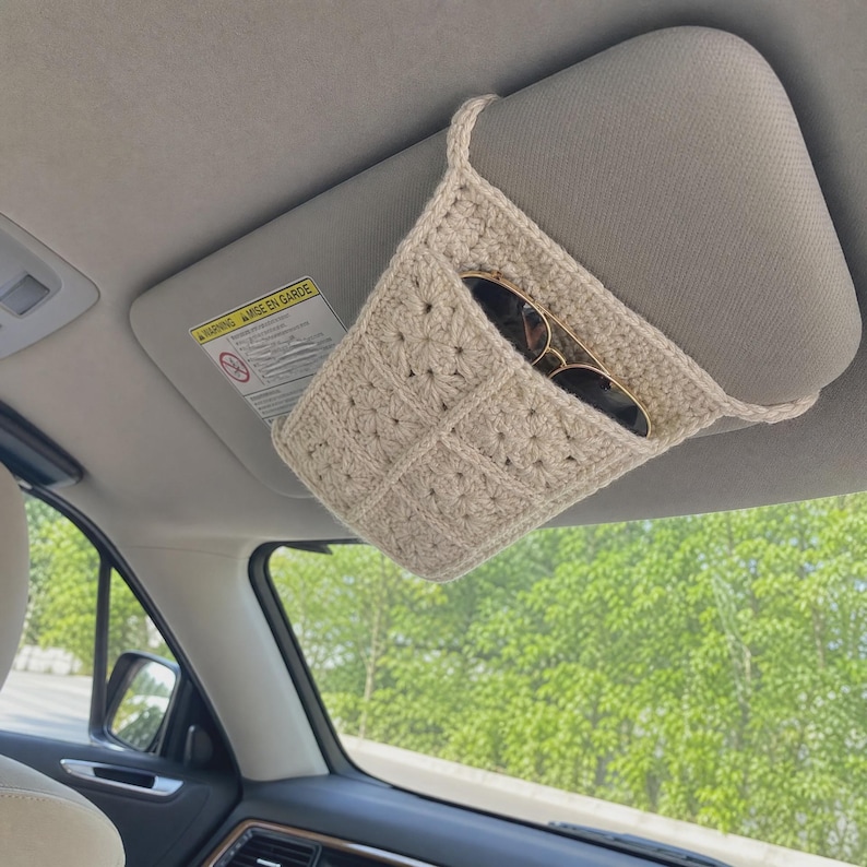 May include: A beige crochet car visor organizer featuring a textured square pattern. The accessory is attached to a vehicle sun visor and contains a pair of dark-rimmed sunglasses tucked into the open pocket, providing storage for small items.