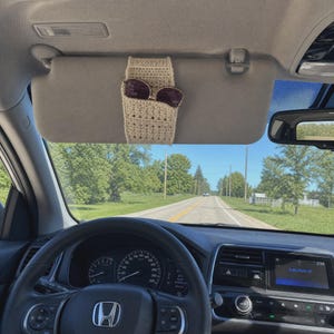 May include: A beige crochet sunglasses holder attached to a car sun visor. The holder features a textured, woven pattern and is designed to securely store a pair of dark-tinted sunglasses, keeping them easily accessible while driving.