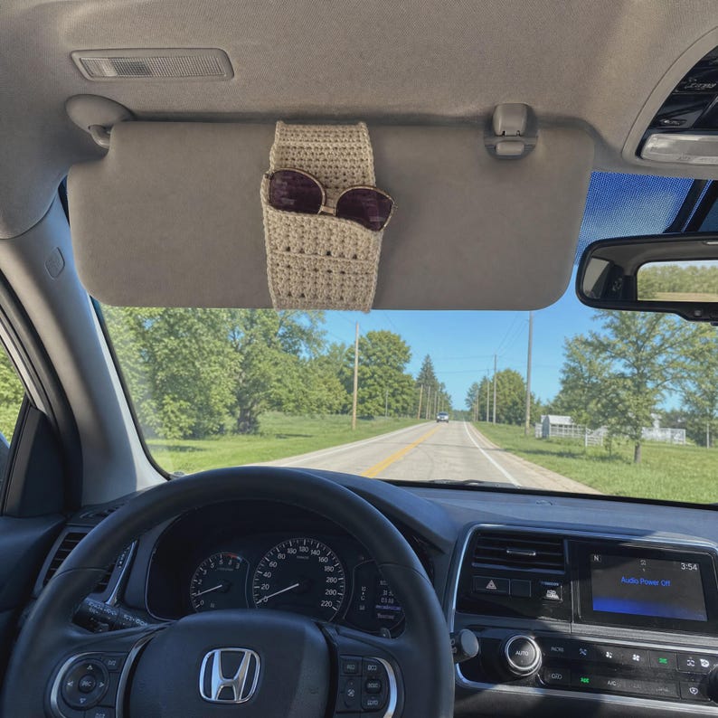 May include: A beige crochet sunglasses holder attached to a car sun visor. The holder features a textured, woven pattern and is designed to securely store a pair of dark-tinted sunglasses, keeping them easily accessible while driving.