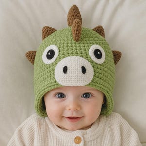 Puede incluir: Un gorro de ganchillo verde con diseño de dinosaurio y púas marrones en la parte superior y los lados. El gorro incluye un hocico blanco con fosas nasales negras y dos ojos grandes blancos con pupilas negras, tejido con punto texturizado.