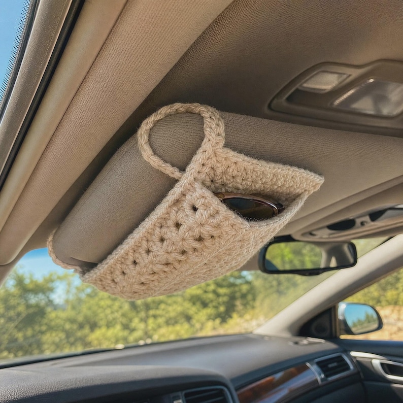May include: A beige crochet car visor organizer featuring a rectangular pouch design with a top loop for attachment. The accessory is mounted on a vehicle sun visor and contains a pair of dark-framed sunglasses tucked inside the open pocket.