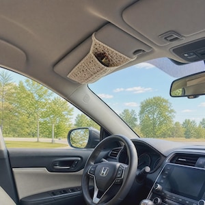 May include: A beige crocheted car visor organizer sleeve attached to the passenger side sun visor. The rectangular fabric sleeve features an open pocket design and is constructed with a textured yarn stitch pattern, holding a pair of sunglasses.