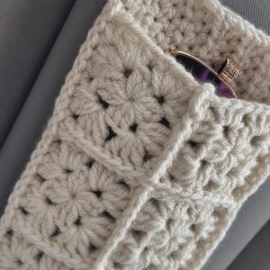 May include: A cream-colored crochet pouch constructed from multiple square motifs with a floral-like pattern. The pouch features an open top edge where a pair of dark-tinted sunglasses with a metallic frame is partially visible inside the sleeve.