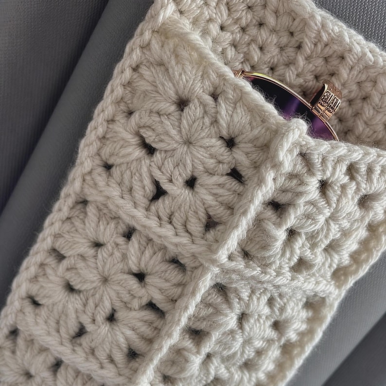 May include: A cream-colored crochet pouch constructed from multiple square motifs with a floral-like pattern. The pouch features an open top edge where a pair of dark-tinted sunglasses with a metallic frame is partially visible inside the sleeve.