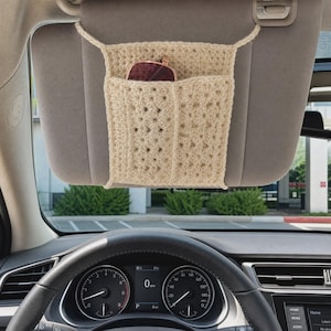 May include: A beige crocheted car visor organizer featuring a rectangular pocket design with a textured stitch pattern. The organizer is attached to a vehicle sun visor and contains a pair of dark-rimmed sunglasses tucked into the front compartment.