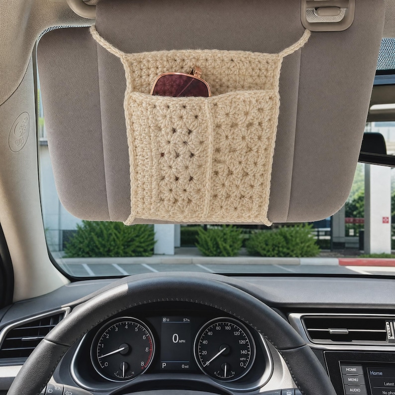 May include: A beige crocheted car visor organizer featuring a rectangular pocket design with a textured stitch pattern. The organizer is attached to a vehicle sun visor and contains a pair of dark-rimmed sunglasses tucked into the front compartment.