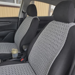 May include: A pair of light gray crocheted seat covers featuring a textured, repeating stitch pattern. These covers are fitted over the front seats of a vehicle, providing a layer of fabric over the black upholstery of the headrests and seats.