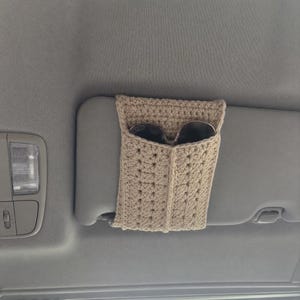 May include: A beige crochet sunglasses holder sleeve attached to a car sun visor. The rectangular holder features a textured stitch pattern and an open top pocket containing a pair of dark-tinted sunglasses with black frames.