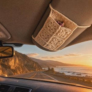 May include: A cream-colored, crocheted fabric organizer sleeve attached to a car sun visor. The sleeve features a textured, grid-like pattern and is designed to hold small items securely against the visor, visible inside a vehicle interior.
