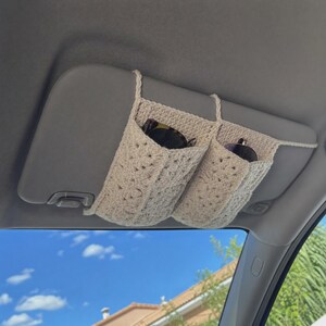 May include: A cream-colored crochet car visor organizer featuring two pockets attached to a vehicle sun visor. The organizer is constructed from yarn with a textured pattern and contains two pairs of sunglasses tucked into the open compartments.