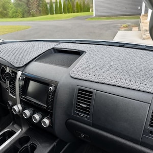 Peut inclure: Une housse de tableau de bord en crochet gris avec un motif de points texturé et répétitif. La housse est ajustée sur la surface supérieure du tableau de bord d'un véhicule, couvrant les côtés conducteur et passager tout en laissant l'aération centrale dégagée.