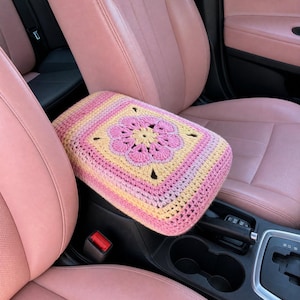 May include: A rectangular crochet armrest cover featuring a central pink and yellow floral motif surrounded by concentric square patterns in shades of pink, yellow, and cream, fitted over the centre console of a vehicle with pink leather seats.