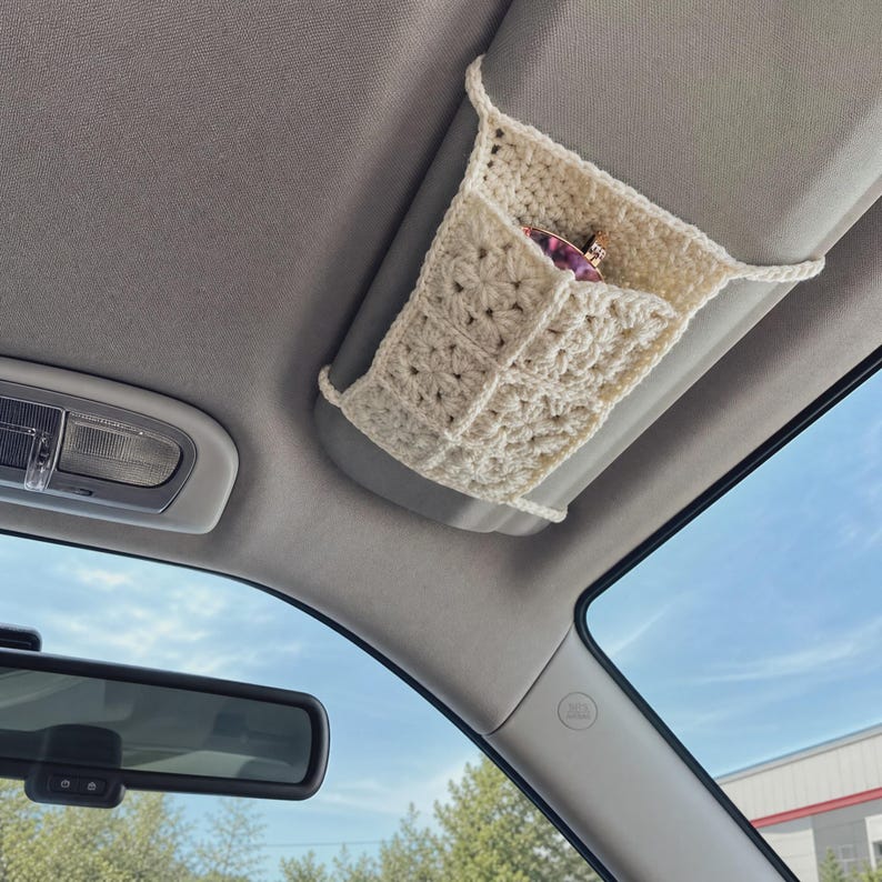 May include: A cream-colored crocheted car visor organizer featuring a square-patterned texture. The rectangular holder is attached to the vehicle's sun visor and contains a pair of sunglasses with dark frames tucked inside the open pocket.