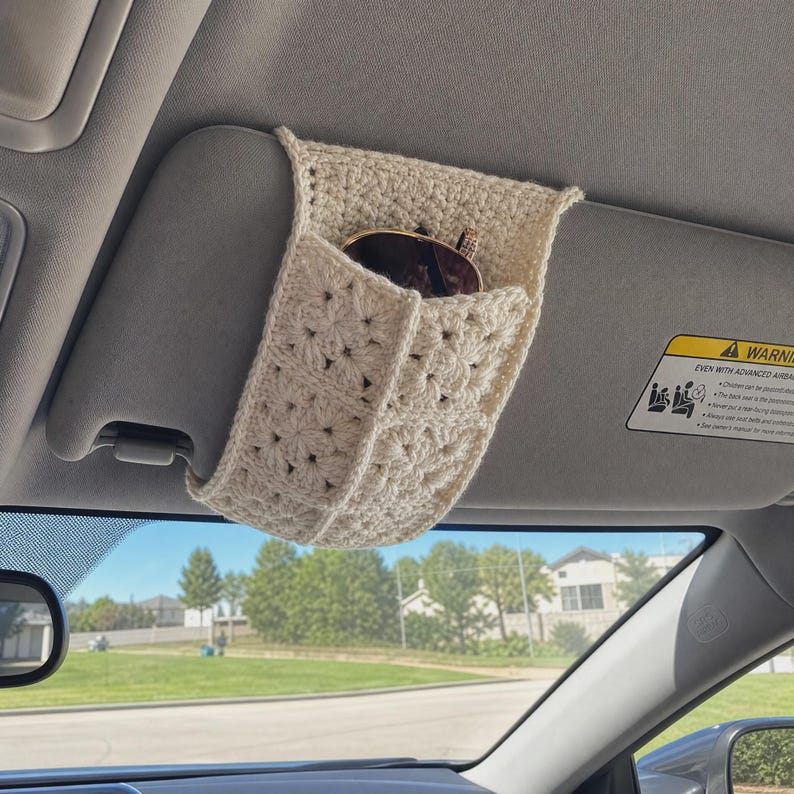 May include: A cream-colored, crocheted car visor organizer featuring a textured granny square pattern. The sleeve is fitted over a vehicle sun visor and contains a pair of dark-rimmed sunglasses tucked into the open pocket compartment.