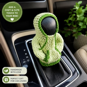 Pode incluir: Uma capa de crochê verde-claro em formato de moletom com capuz para a alavanca de câmbio de um carro. A peça em miniatura possui capuz, mangas longas e um bolso frontal, com acabamento em verde-escuro nas bordas do capuz, punhos e bainha.