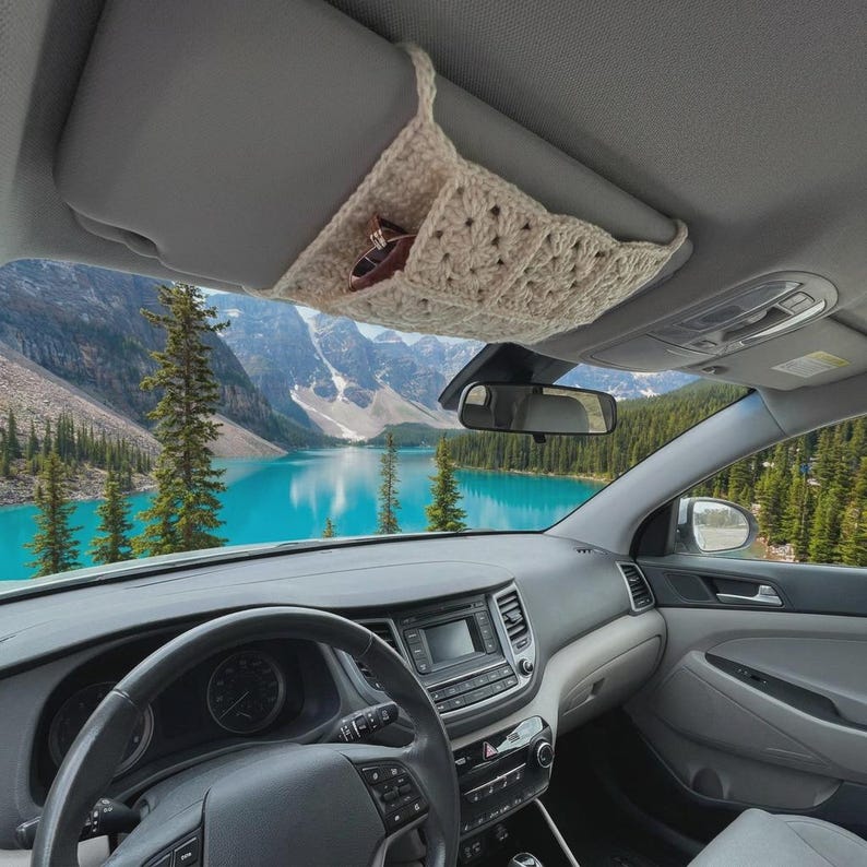 May include: A cream-colored crocheted organizer sleeve attached to a car sun visor. The sleeve features a square-patterned knit design and contains a pair of dark-rimmed sunglasses tucked into one of the open pockets for storage and accessibility.