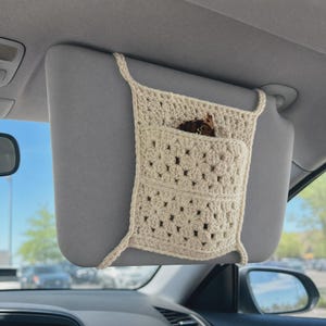 May include: A cream-colored crochet car visor organizer featuring a square granny square pattern with a central pocket. The organizer is secured to a gray vehicle sun visor with crocheted straps and contains a pair of brown sunglasses.